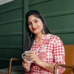 Positive smiling Indian lady in stylish traditional wear sitting on chair against green wall with cup of hot drink and looking at camera